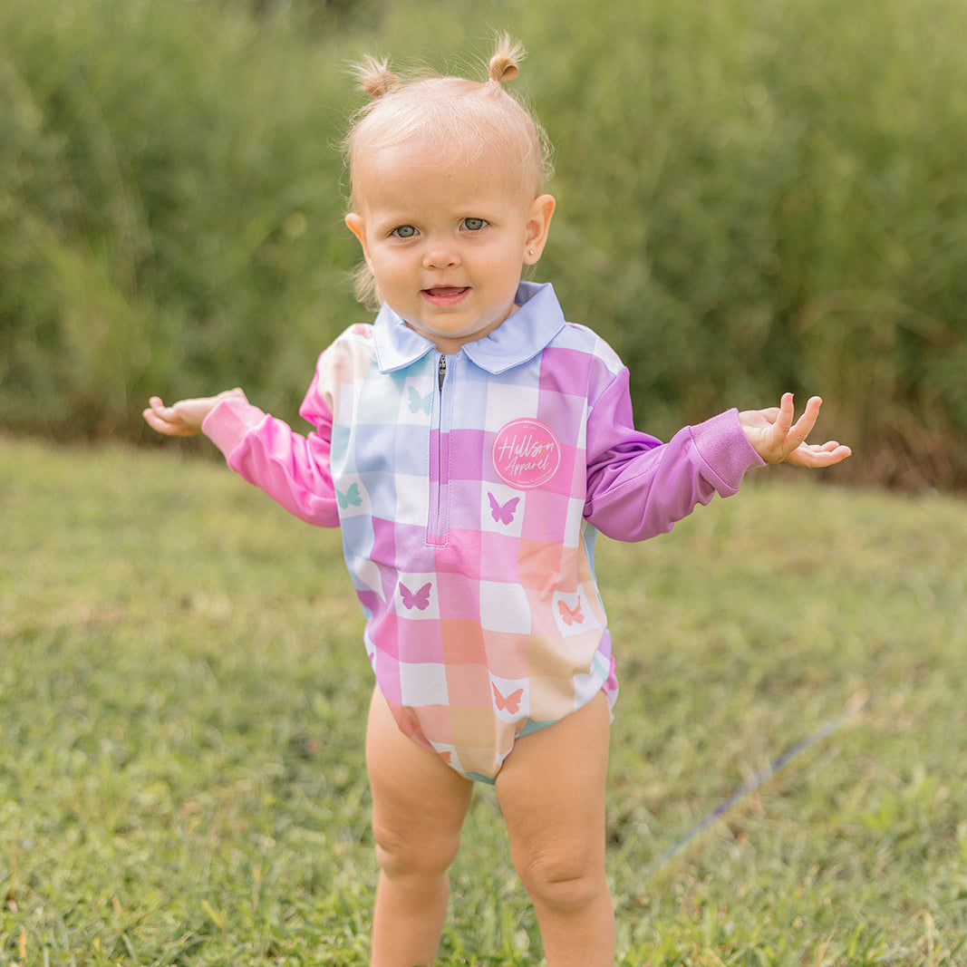 Baby in a colorful fishing romper standing on grass with trees in the background