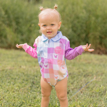 Baby in a colorful fishing romper standing on grass with trees in the background