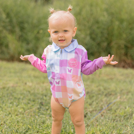 Baby in a colorful fishing romper standing on grass with trees in the background