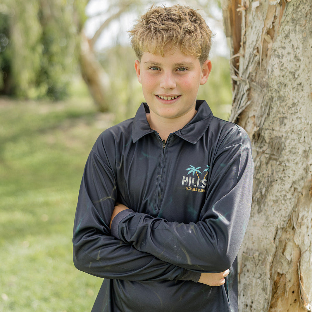 Young boy wearing a Hillson apparel fishing shirt leaning against a tree outdoors.