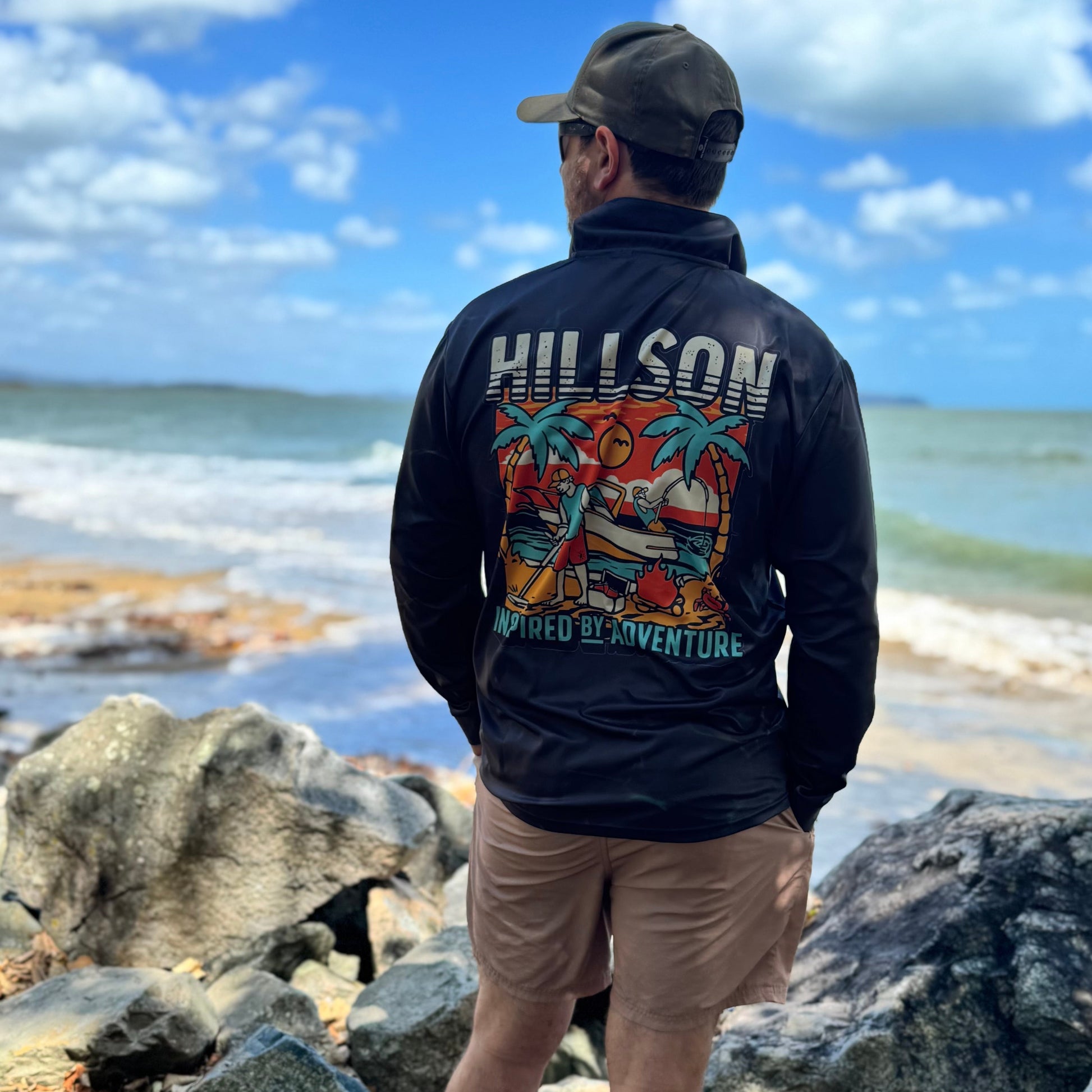 Person wearing a black jacket with colorful designs and text, standing on rocks by the ocean.