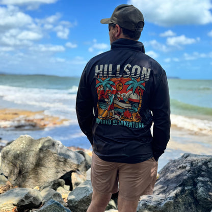 Person wearing a black jacket with colorful designs and text, standing on rocks by the ocean.